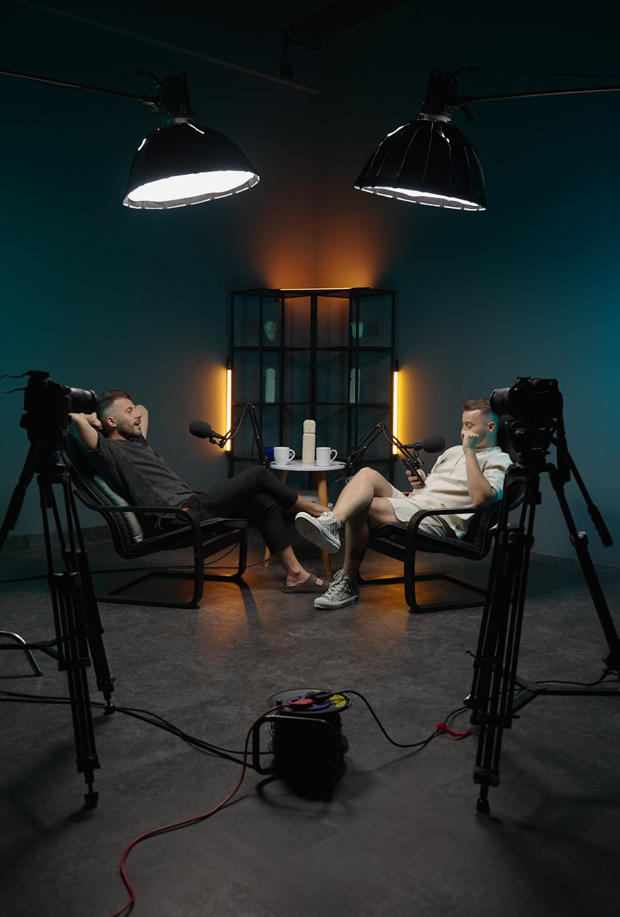 Two speakers in armchairs recording a conversation under studio lights